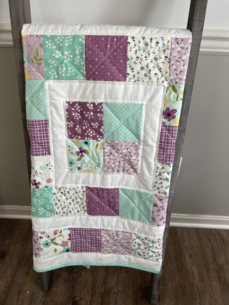 Image contains a quilt made from white, teal, and purple fabric squares, displayed on a wooden ladder.