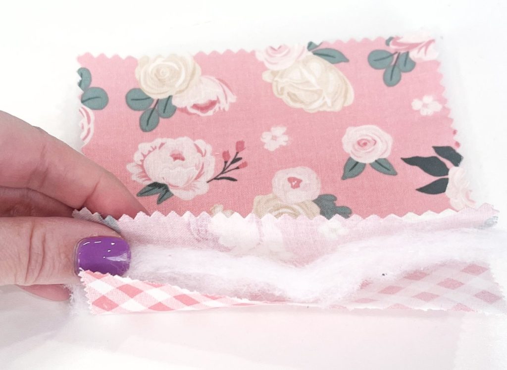 Image contains Amy’s hand holding a stack of two 5" pieces of fabric with a 5” piece of batting in between.
