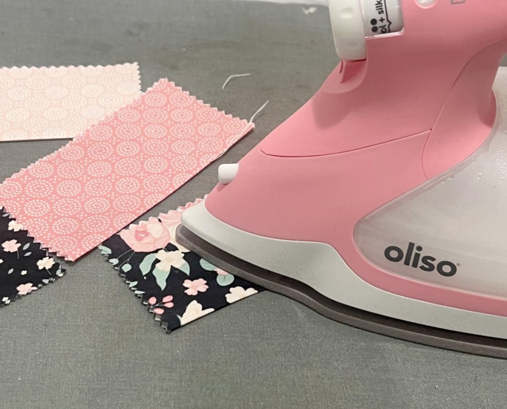 Image contains a pink oliso iron pressing 5” fabric squares in half.