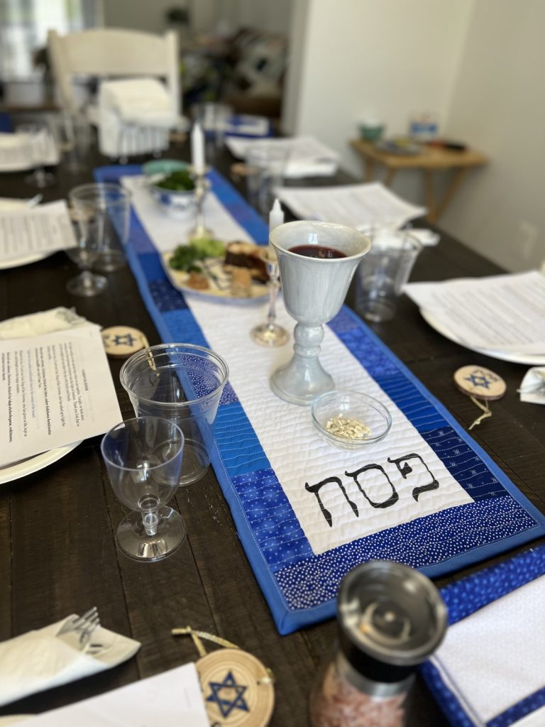Image contains a wooden-topped table set for Passover Seder, with a white and blue table runner in the center.