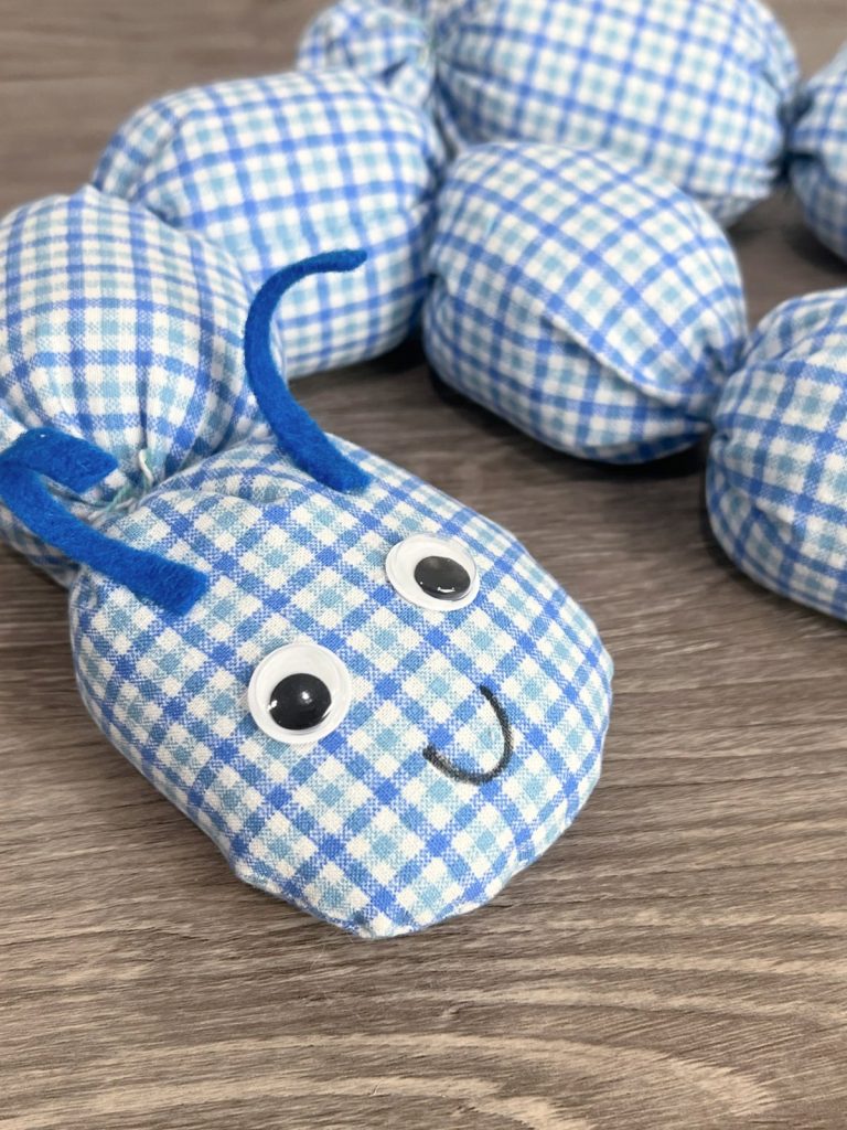 Image contains a close-up view of a plush caterpillar made from blue and white plaid fabric, with google eyes, blue felt antennae, and a small black smile.