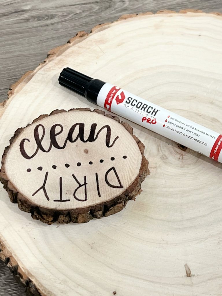 Image shows a wood burned project; a small wood slice with the words “clean” and dirty” burned onto it. It sits on top of a larger wood slice, with a marker nearby.