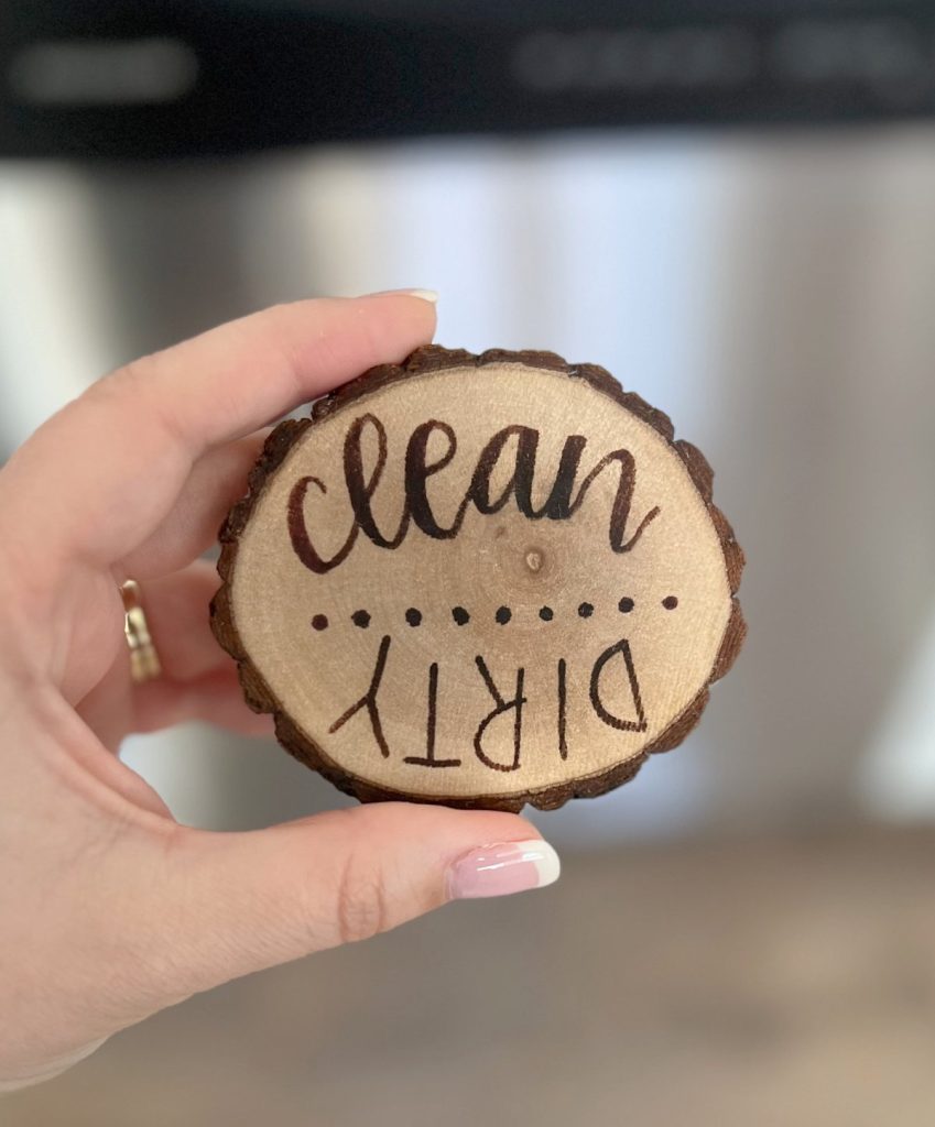 Image contains Amy’s hand holding a small wood slice magnet with the words “clean” and “dirty” wood burned onto it.