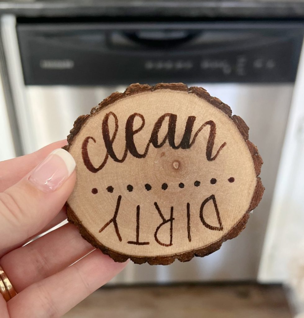 Image contains Amy’s hand holding a wood slice magnet with the words “clean” and “dirty” wood-burned onto it.