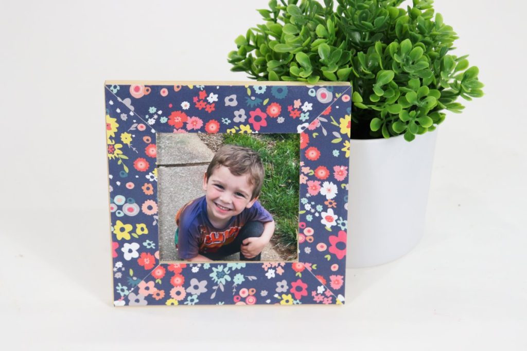 Image contains a blue floral decoupaged photo frame with a picture of a smiling male child in the center. A faux plant sits behind the frame on a white table.