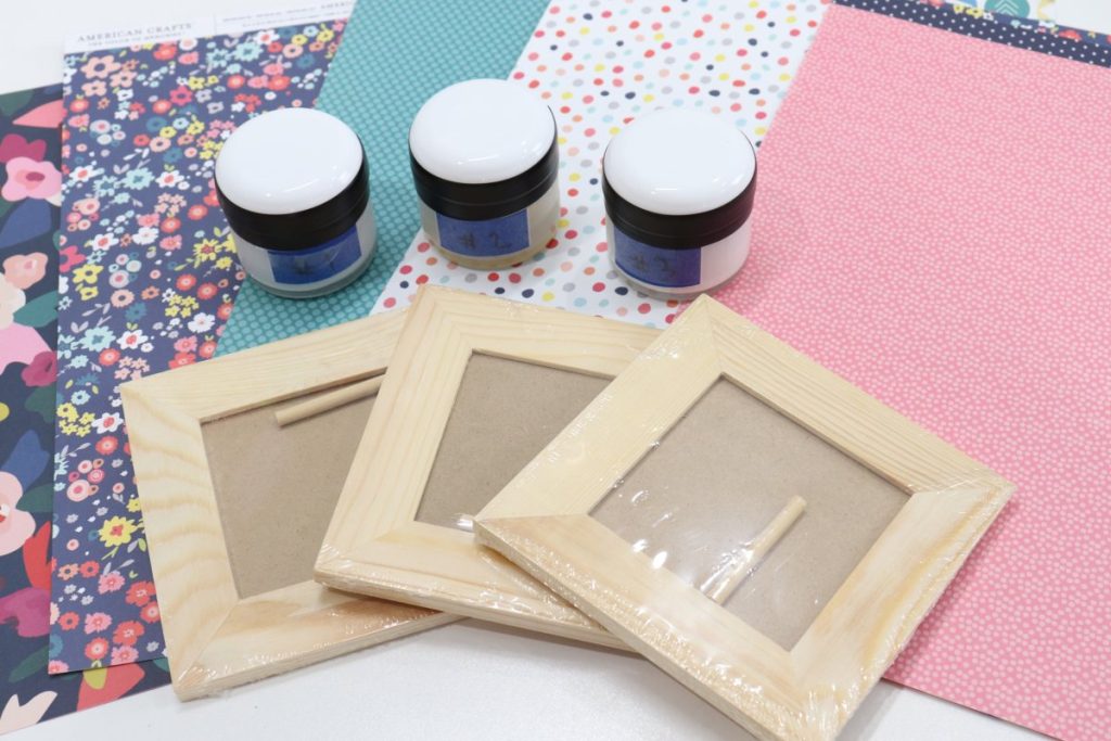 Image contains three unfinished wooden photo frames, three jars of decoupage formula, and assorted scrapbook paper in shades of pink, teal, and multicolored.