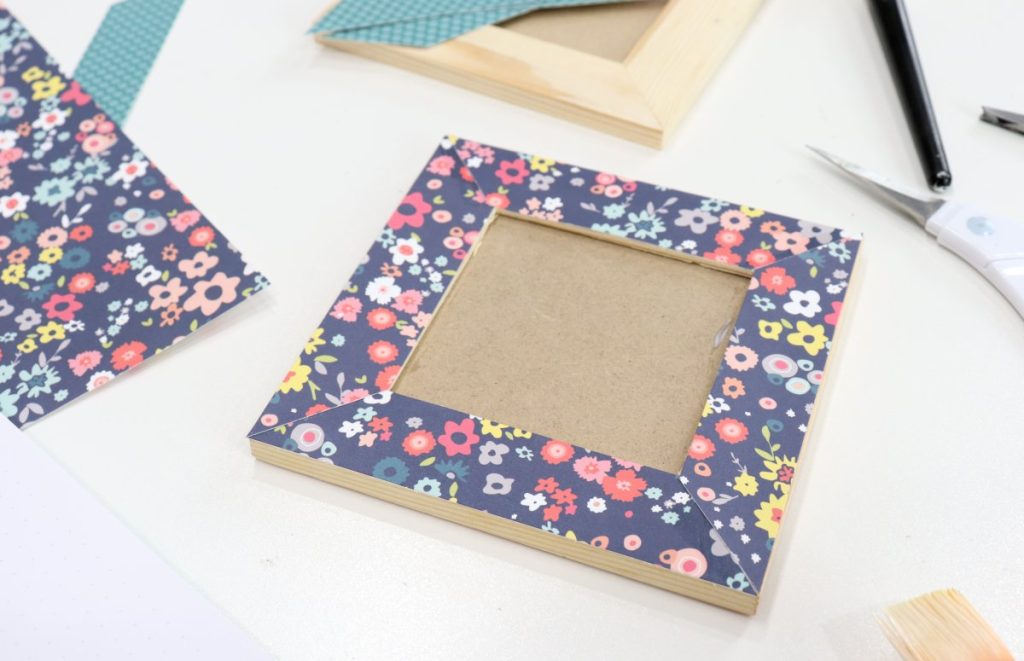 Image contains a square wooden photo frame with blue floral paper decoupaged to it. Assorted craft supplies, like scissors and other scrapbook paper and frames are scattered in the background.