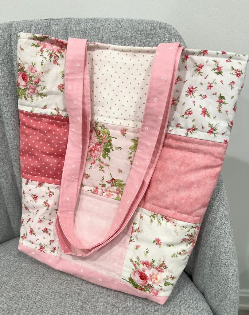 Image contains a fabric tote bag with pink, white, and floral patchwork squares and pink handles.