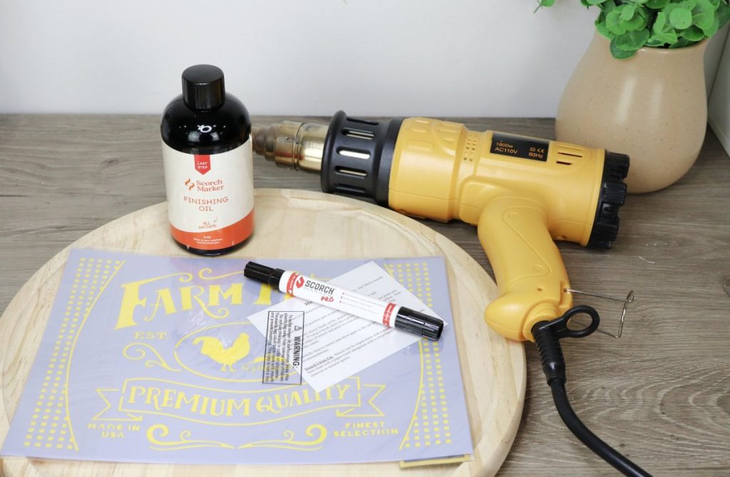 Image contains the project supplies: an unfinished round wood tray, a farmhouse stencil, wood finishing oil, the Scorch marker Pro, and a yellow heat tool.