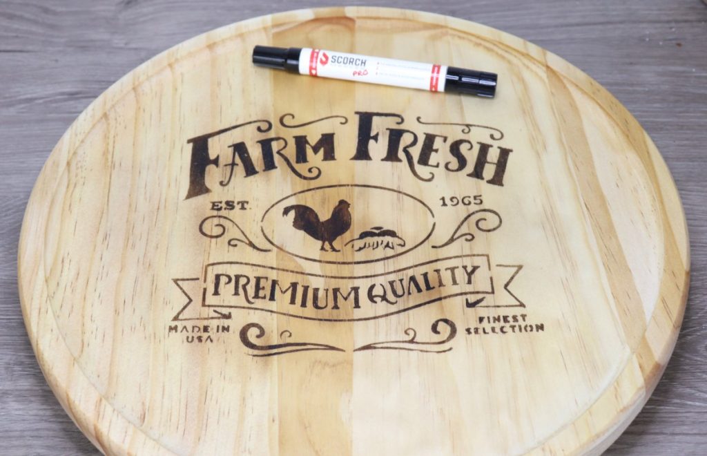 Image contains a round wooden tray with a “farm fresh” farmhouse design wood-burned onto the surface. A Scorch Marker sits on the tray.