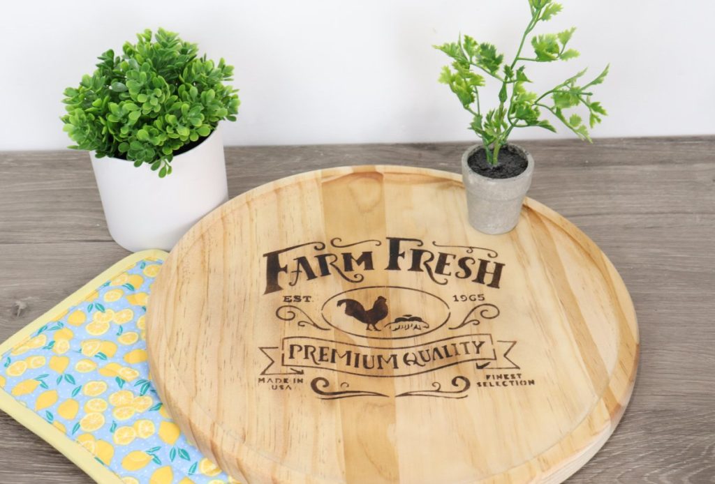 Image contains a round wooden tray with a “farm fresh” farmhouse design wood-burned onto the surface. Faux plants sit on and around the tray, and there is a blue pot holder with yellow lemons to the left.