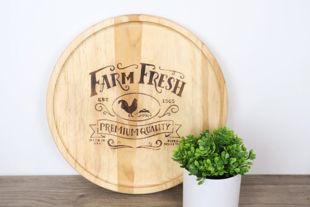 Image contains a round wooden tray with a “farm fresh” farmhouse design wood-burned onto the surface.