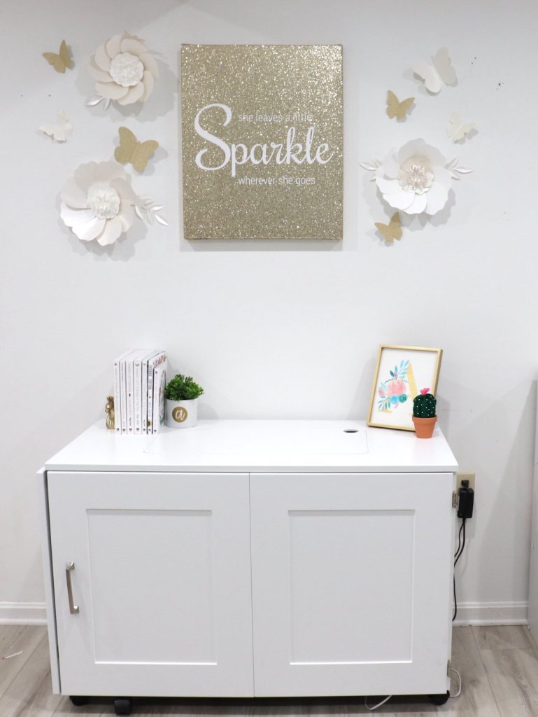 Image contains the Sew Station folded into a small cabinet with white and gold decorations on the wall above it.