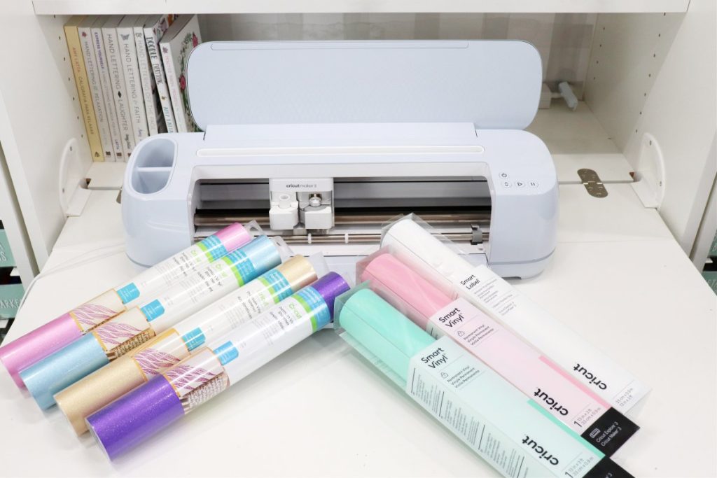 Image contains a light blue Cricut Maker 3 machine surrounded by rolls of Shimmer Vinyl and Smart Vinyl.