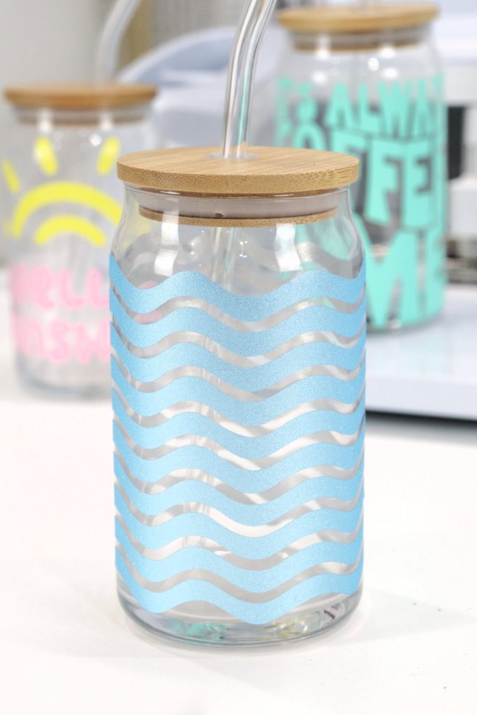 Image contains a glass cup with a bamboo lid, decorated with blue shimmer vinyl in a pattern of repeating waves. Behind it, out of focus, are two other decorated glasses.