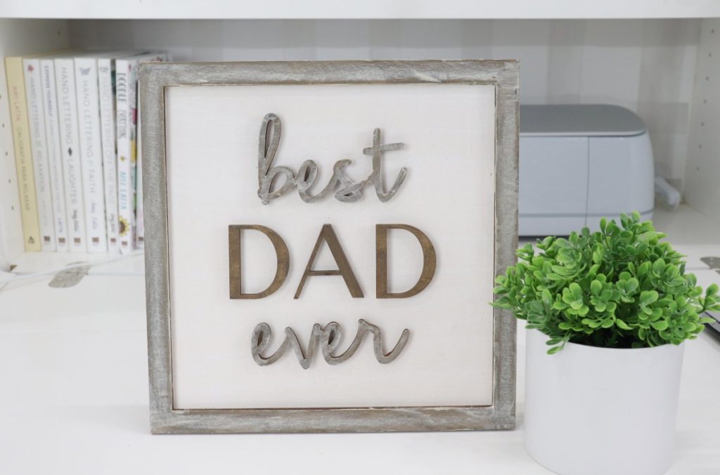 Image contains a wooden sign with a white background, brown frame, and brown words reading, “best dad ever."