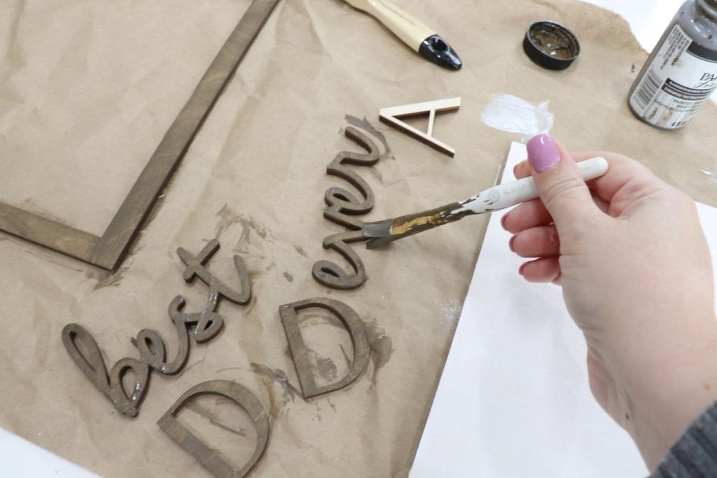 Image contains Amy’s hand holding a paintbrush and applying wood stain to cutout words and letters.