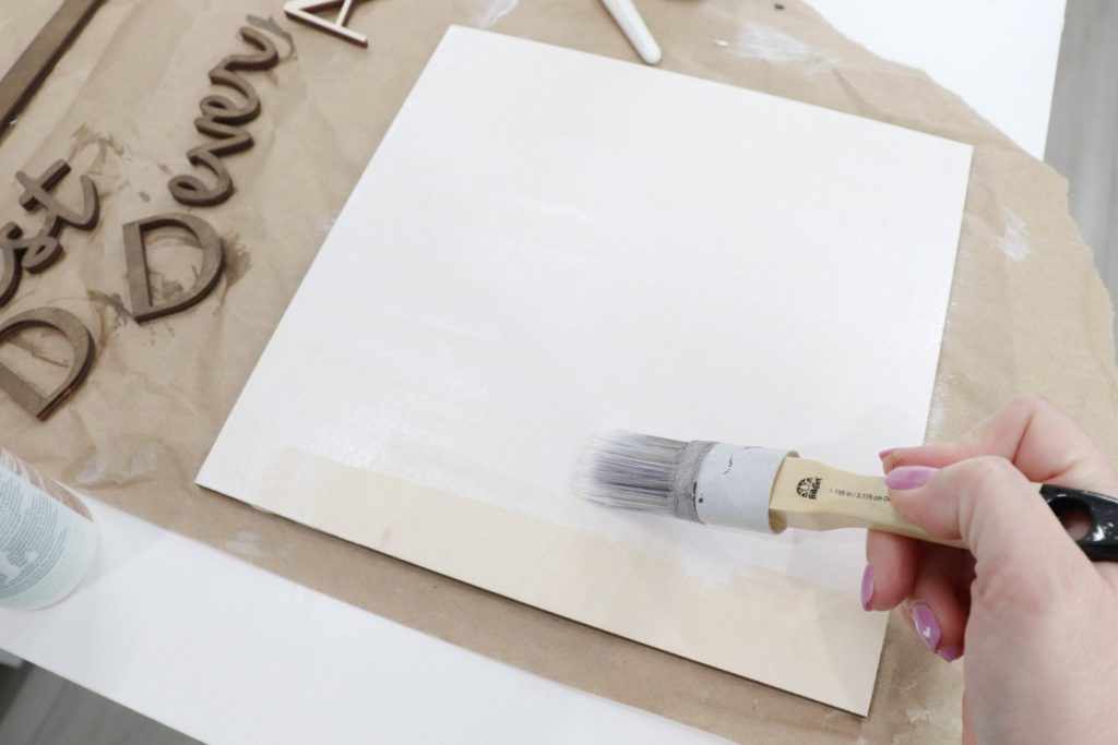 Image contains Amy’s hand holding a paintbrush and applying white wood stain to a wooden square.