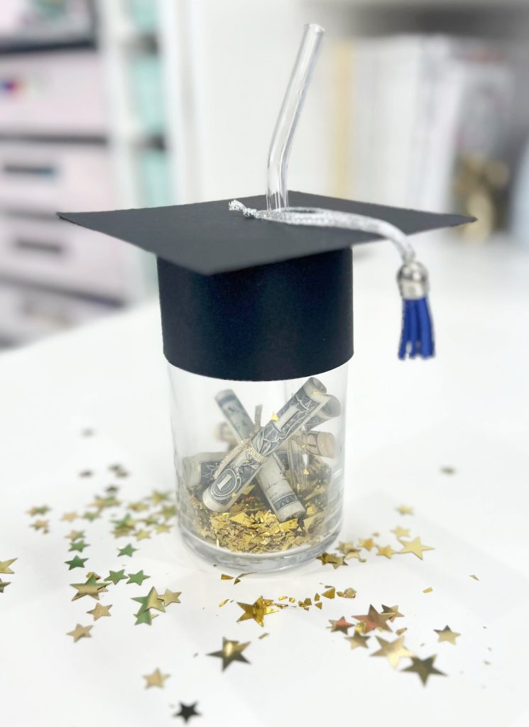 Image contains a glass cup filled with confetti and money. The top is decorated to look like a graduation cap made from black cardstock.