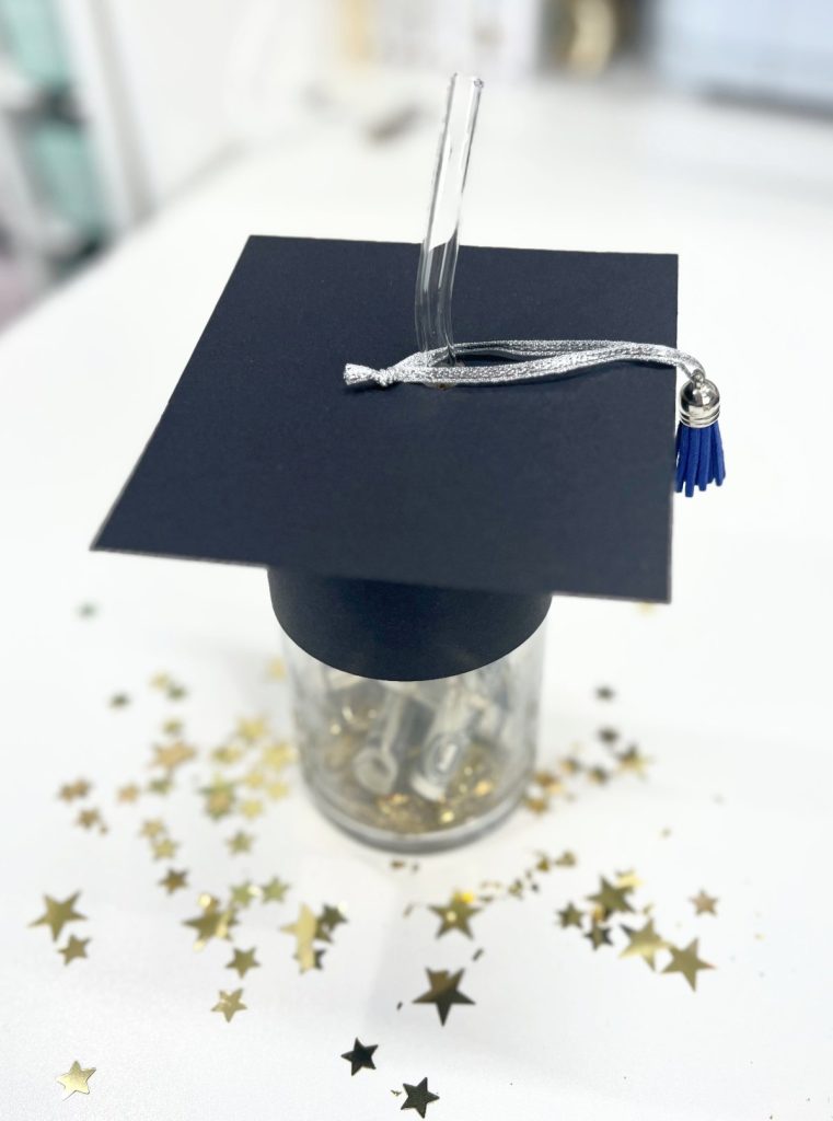 Image contains a glass cup with a black cardstock graduation cap on the lid. It sits on a white table surrounded by gold star confetti.