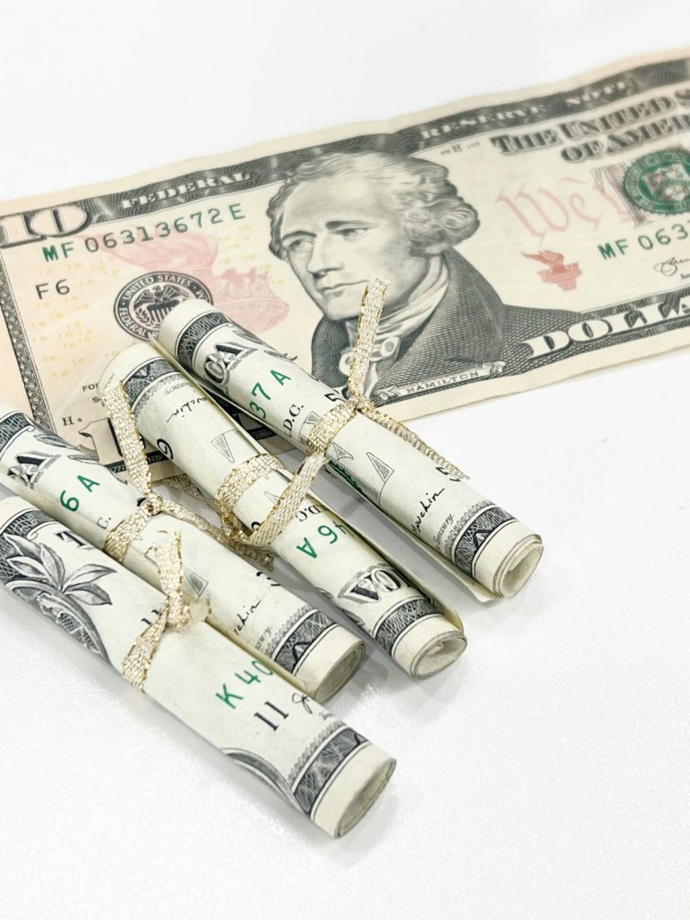 Image contains a ten dollar bill laying flat on a white table with rolled up dollar bills beside it.