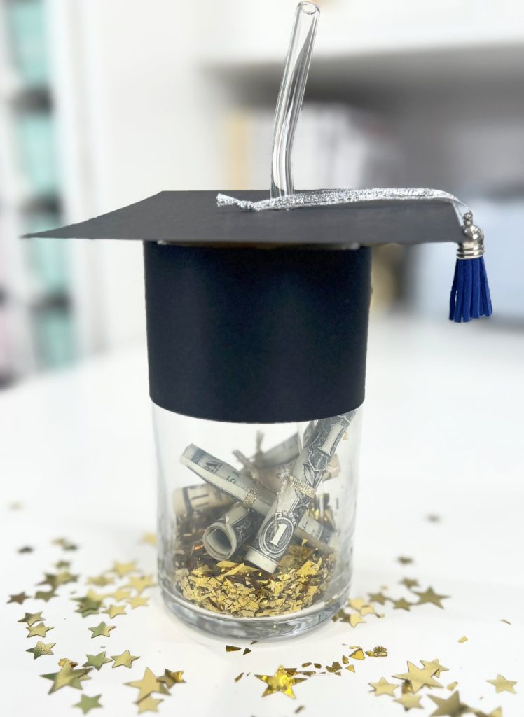 Image contains a class cup with a black cardstock graudation cap on top. The cup is filled with gold confetti and rolled up money.