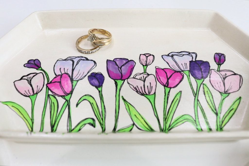 Image contains a white jewelry tray decorated with tulips in varying shades of purple and pink. A pair of gold rings sits on the tray.