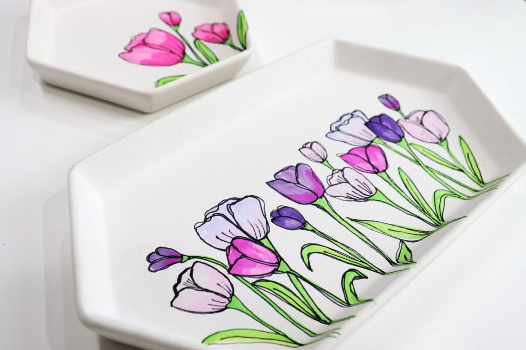 Image contains two white jewelry trays with pink and purple tulips drawn on them using alcohol marker.