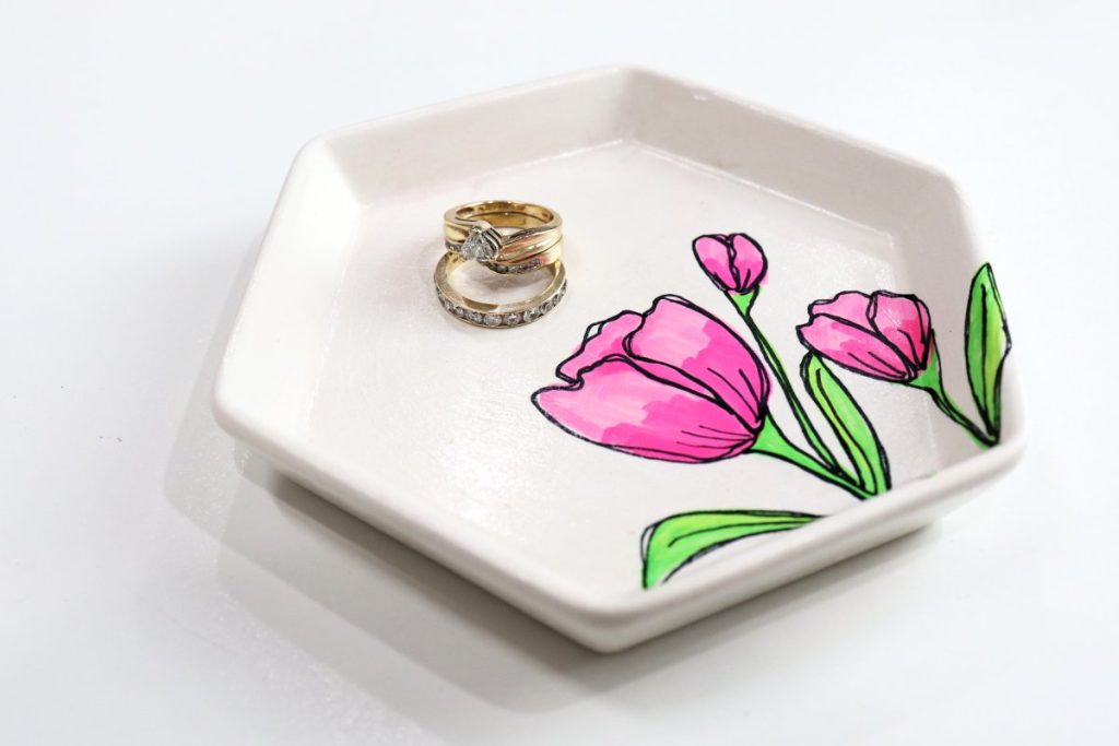 Image contains a white jewelry tray with three pink tulips drawn on it. Two gold rings sit in the tray.