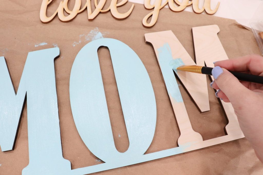 Image contains Amy’s hand painting the wooden word “MOM” with teal paint.