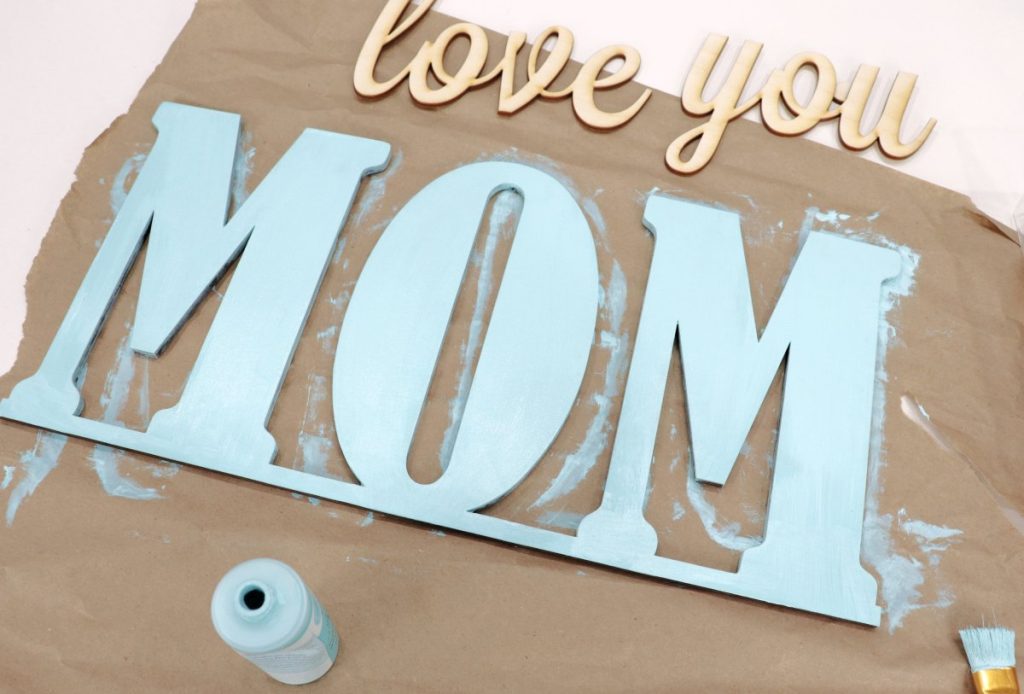 Image contains the words, “love you mom,” cut out of light wood. The “mom” is painted teal.