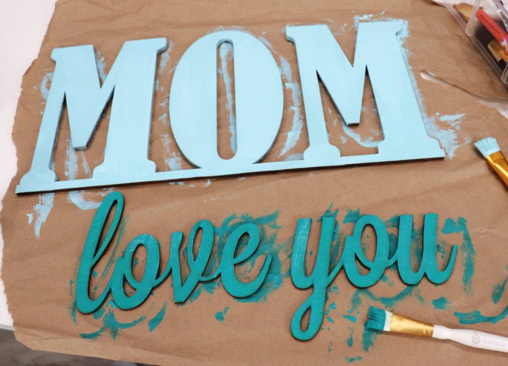 Image contains the words “mom” and “love you” cut from wood. “Mom” is painted a light teal, and “love you” is painted with a darker teal. Paintbrushes sit nearby.