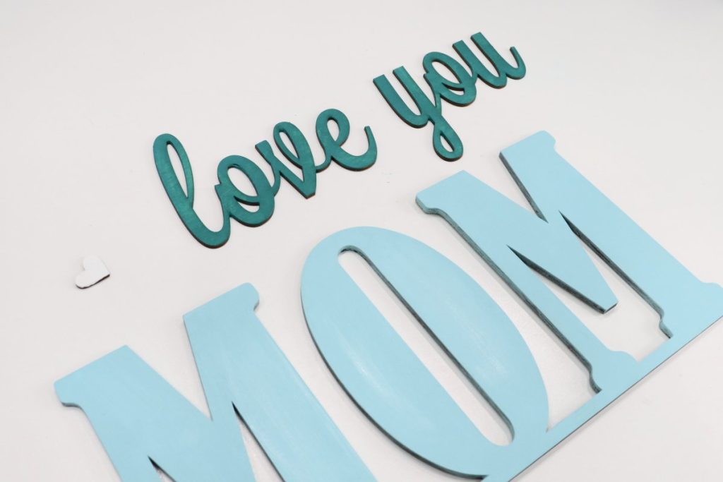Image contains the words, “love you Mom” cut from wood and painted in shades of teal.