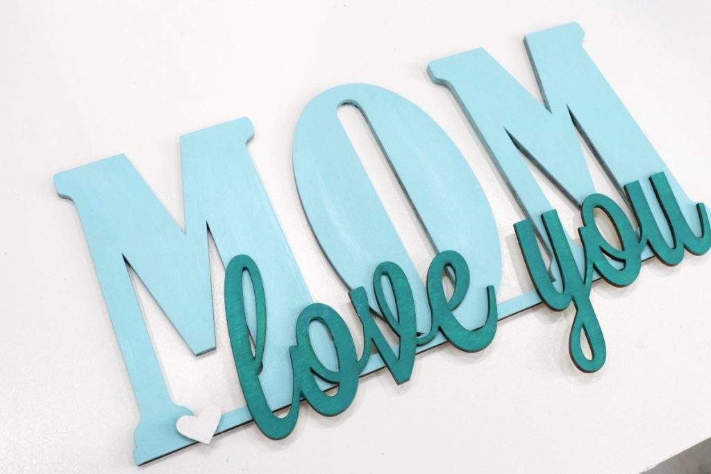 Image contains the words, “love you mom,” cut from wood and painted teal.