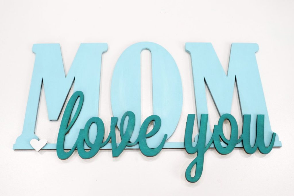 Image contains wooden words, “love you Mom” painted in shades of teal.