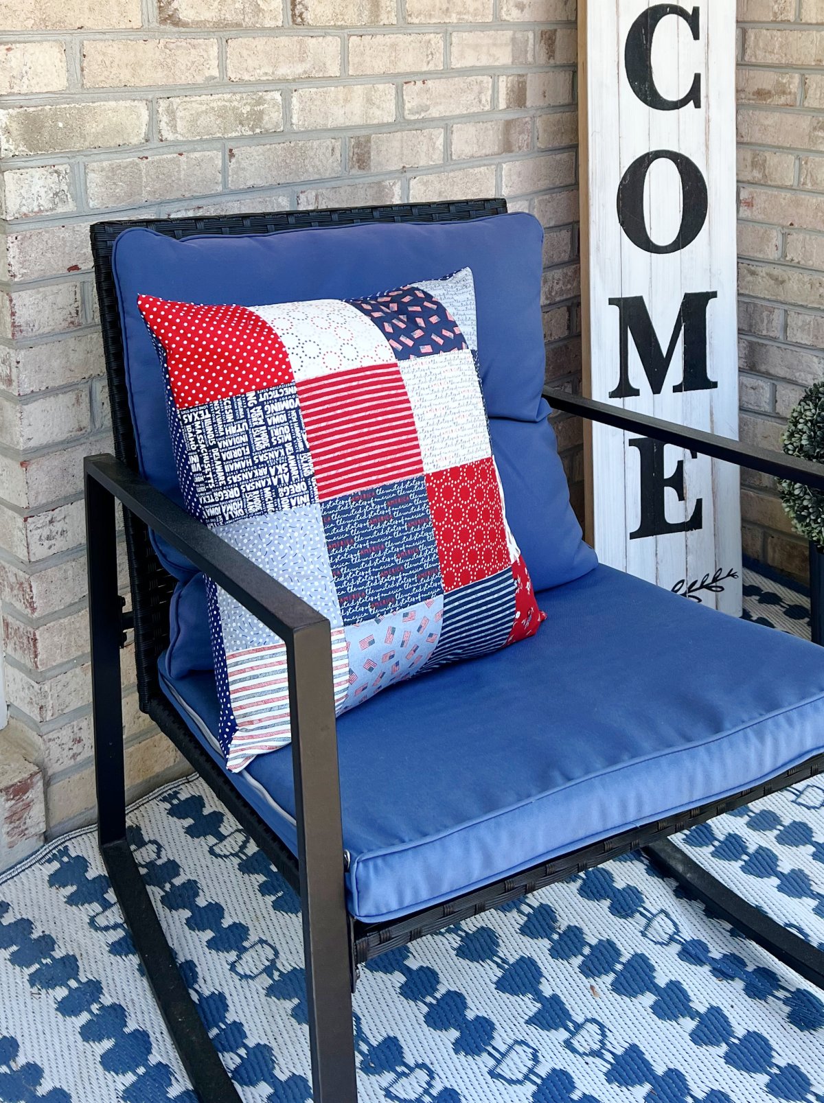 Image contains a square patchwork pillow made from red, white, and blue fabric sitting on a blue outdoor rocking chair.