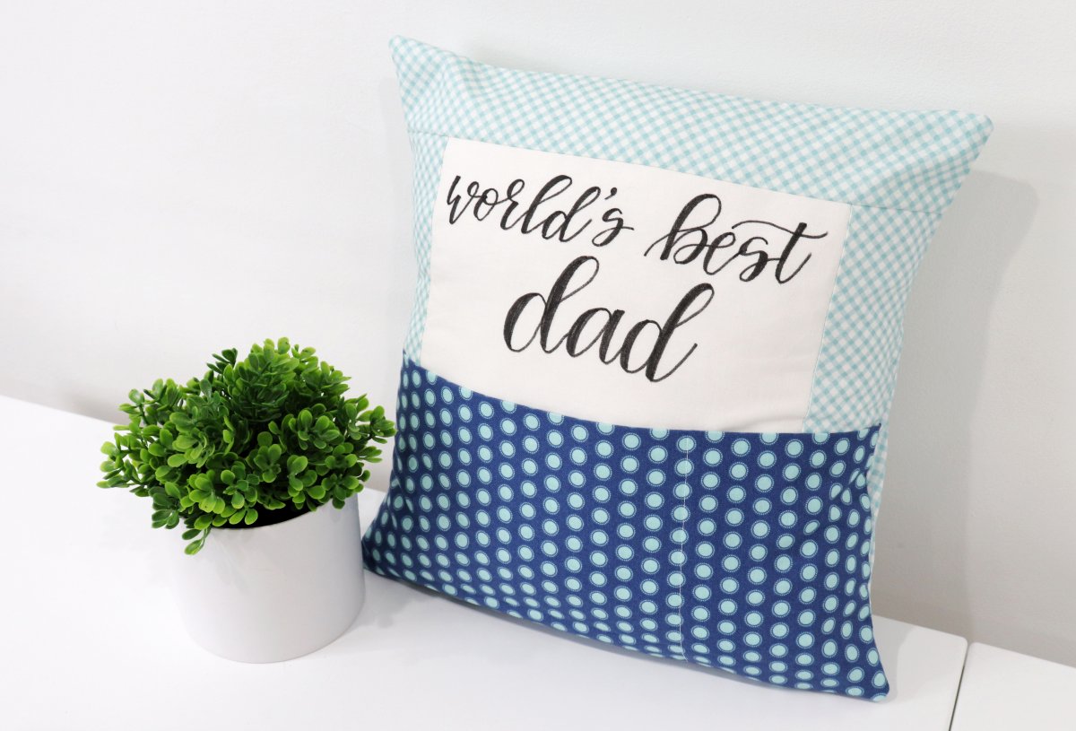 Image contains a pillow cover with a white center, light blue and white sashing, and a dark blue print pocket. The words “world’s best dad” are lettered in black fabric marker.