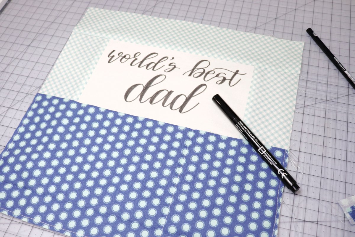 Image contains a fabric pillow cover front with the words “world’s best dad” hand lettered on it in black. A fabric marker sits on top.