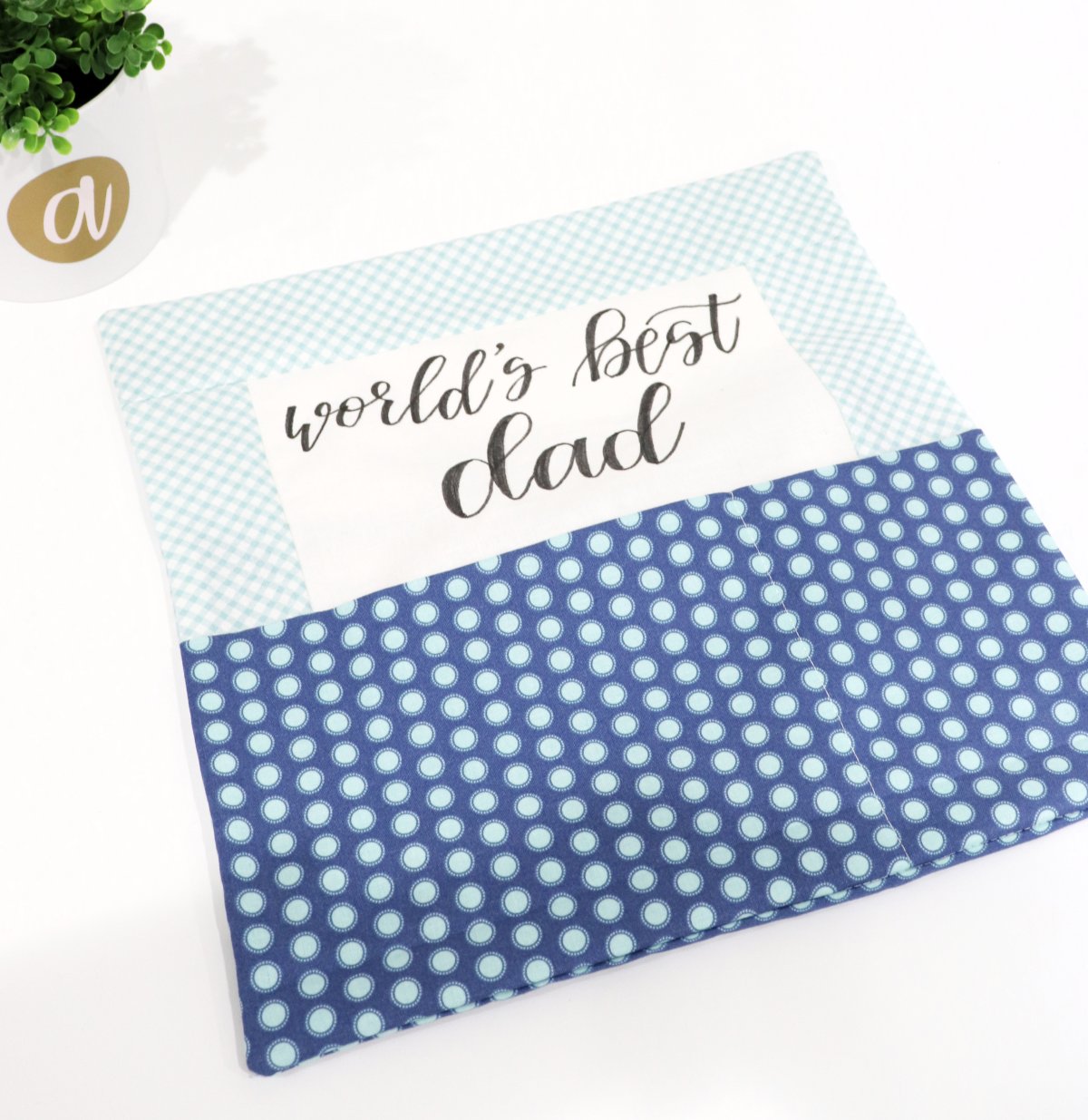 Image contains a pillow cover with a white center, light blue and white sashing, and a dark blue print pocket. The words “world’s best dad” are lettered in black fabric marker.