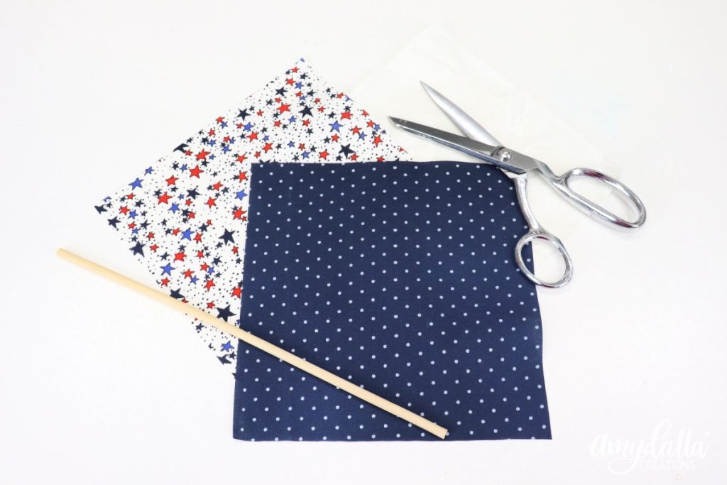 Image contains two fabric squares (one white with colored stars, one blue with white dots), a square of interfacing, a pair of silver scissors, and a wooden dowel on a white table.