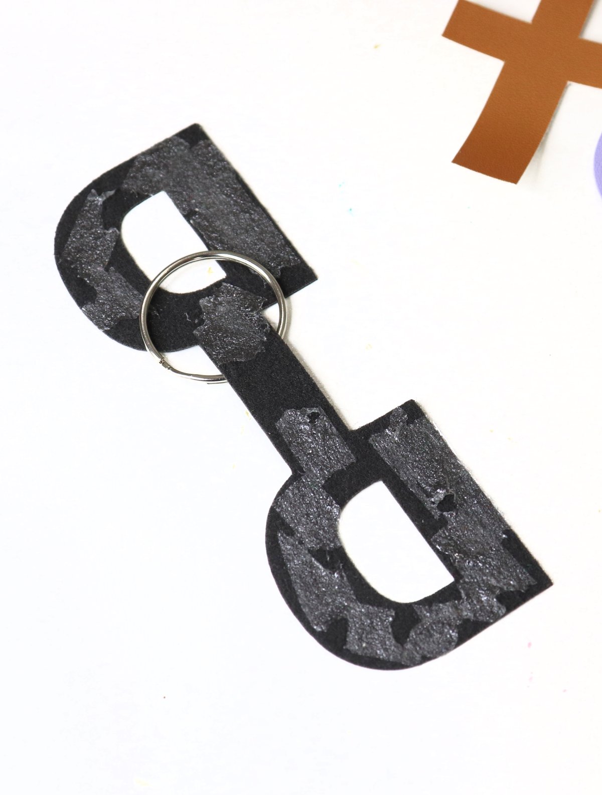 Image contains a black “D” monogram with adhesive on both sides and a keyring in the center.
