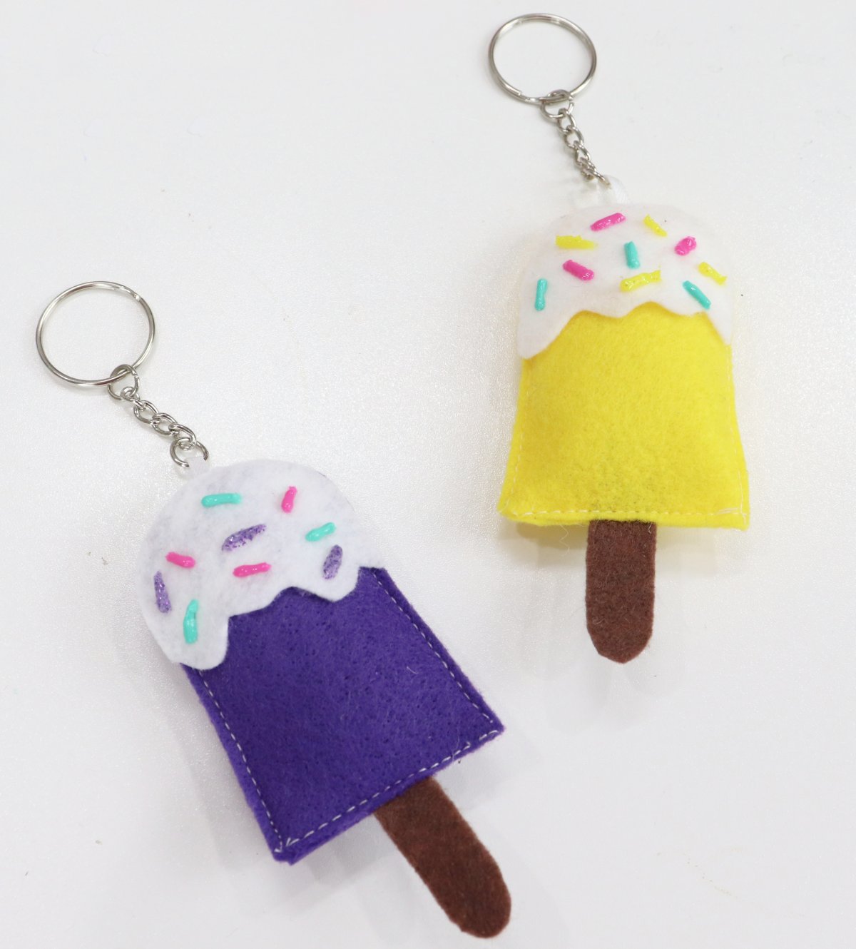 Image contains two felt popsicle keychains; one purple and one yellow, on a white table.