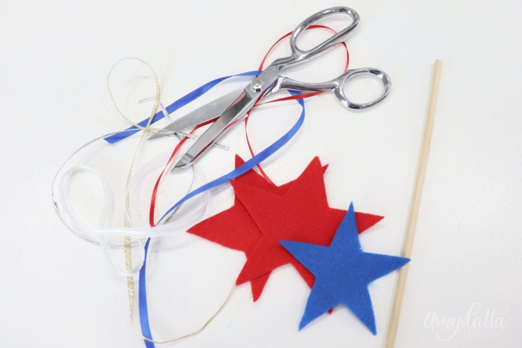 Image contains two red felt stars, one blue star, assorted ribbons, a wooden dowel, and a pair of silver scissors.