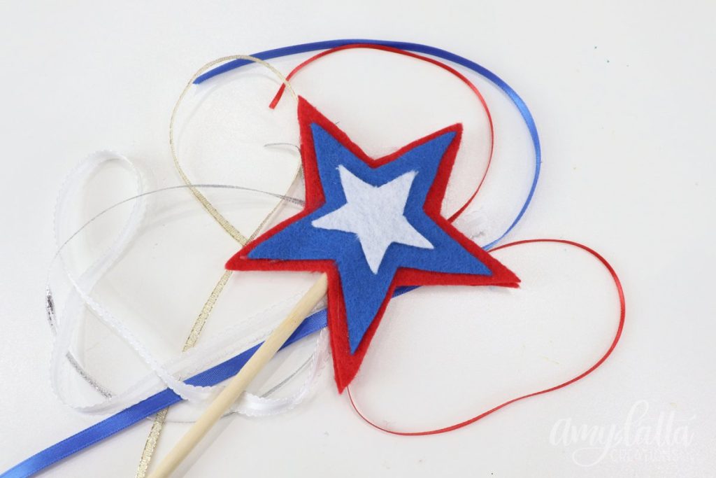 Image contains a wooden dowel with a red, white, and blue felt star topper, sitting on top of red, white, and blue ribbons.