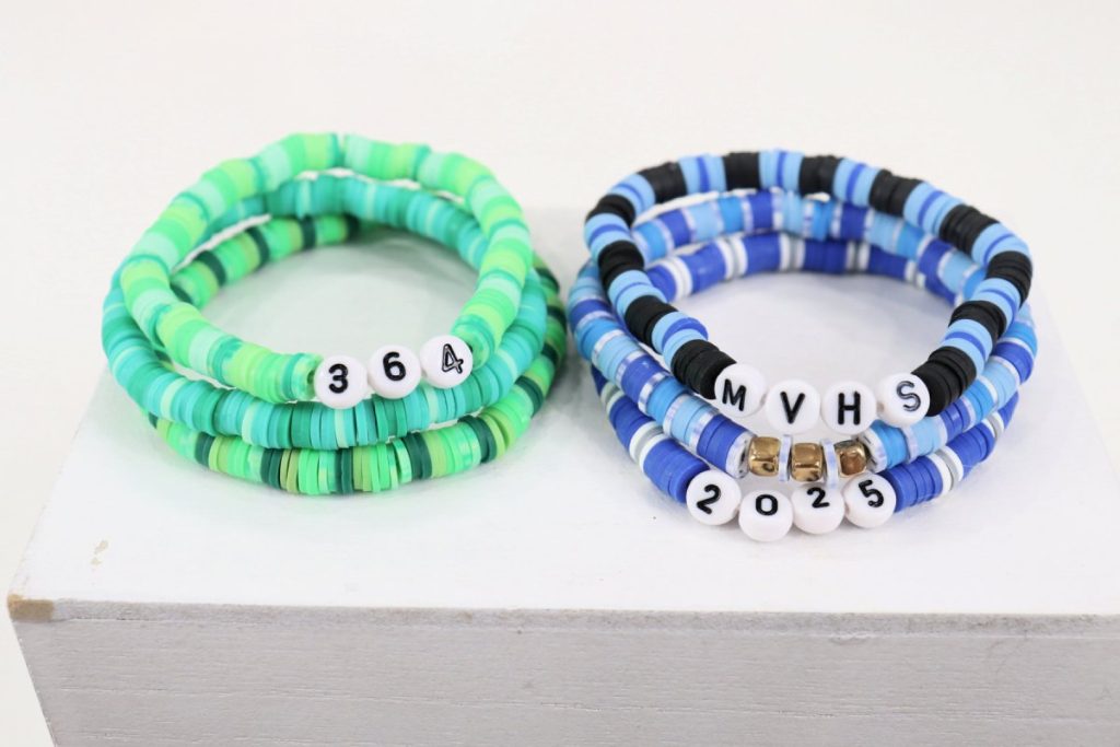 Image contains a stack of three beaded bracelets made from various shades of green clay beads, and a stack of three beaded bracelets made from blue, white, and black clay beads.