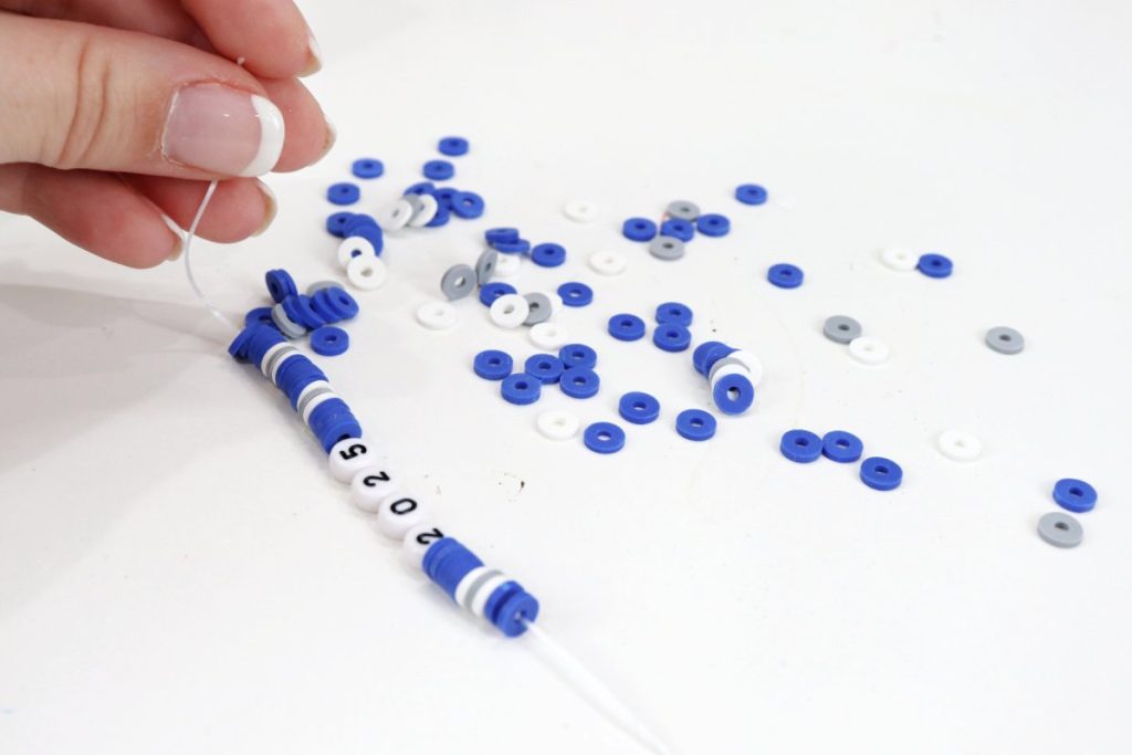 Image contains Amy’s hand holding one end of an elastic cord with blue, white, and grey beads on it. There are more blue, white, and grey beads beside it on a white table.