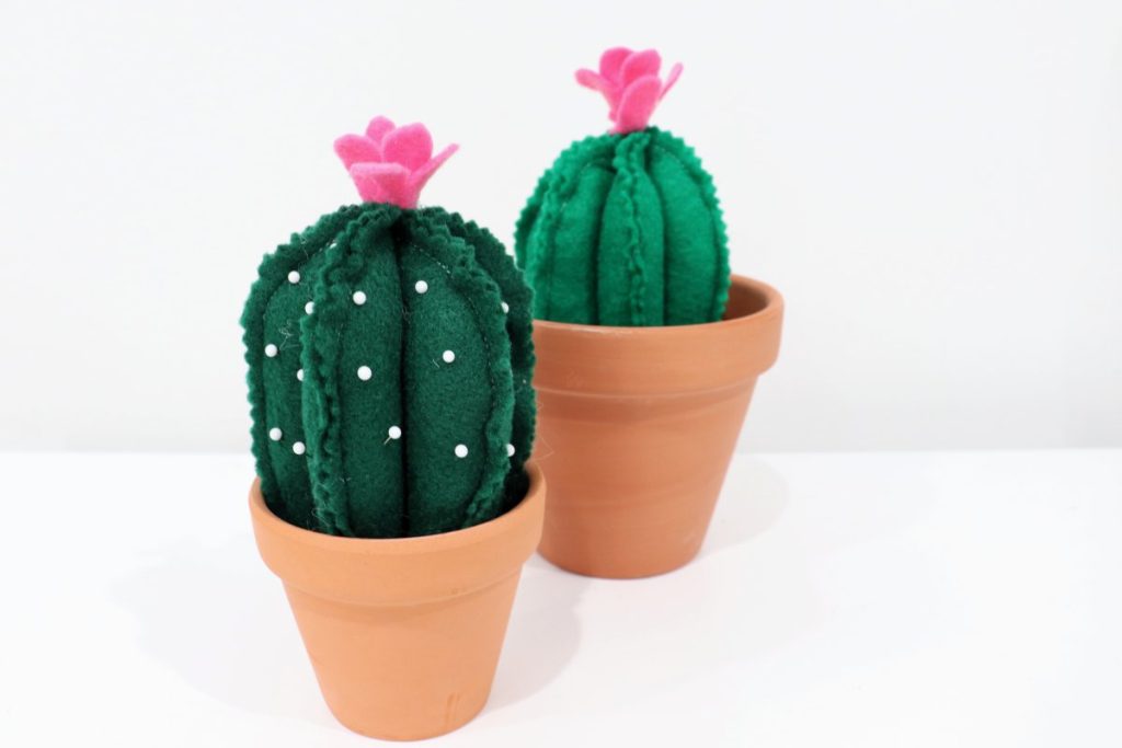 Image contains two decorative cacti made from green felt with pink felt flowers on top, in terracotta pots.