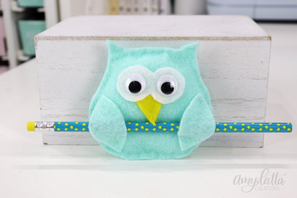 Image contains a teal felt owl holding a turquoise pencil with yellow stars. It is leaning against a white box on a white table.