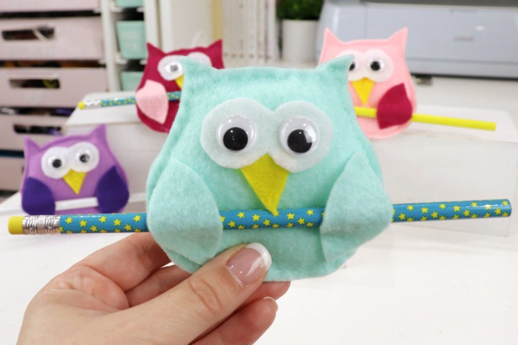 Image contains Amy’s hand holding a teal felt owl with a turquoise and yellow pencil in its wings. Three other felt owls are in the background.
