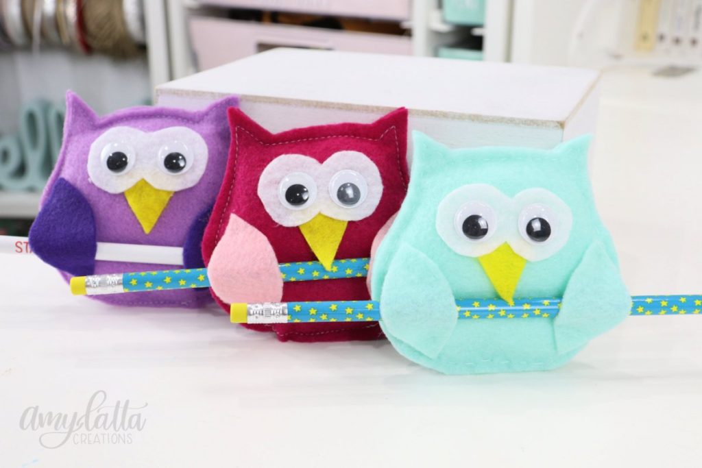 Image contains three felt owls; one purple, one pink, and one light teal, all holding pencils.